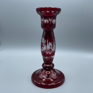 May include: A ruby red glass candlestick with etched white designs. The candlestick features a wide base, a central stem with a detailed etched design, and a flared top. The etched design includes floral and architectural elements.