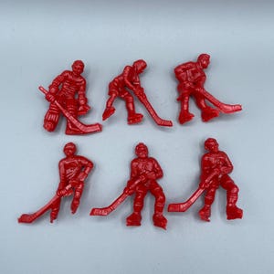 Op de afbeelding: Zeven kleine, rode plastic hockey speler beeldjes. Elke figuur staat in een andere pose, met een hockeystick. De beeldjes zijn allemaal even groot en van dezelfde kleur, tegen een effen witte achtergrond.