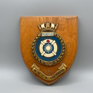 Può includere: Targa in legno a forma di scudo, con un stemma con le scritte "PRESERVER" e "H.M.C.S. PRESERVER". Lo stemma presenta un design blu e oro con un simbolo di salvagente e una corona. La targa è in legno marrone chiaro.