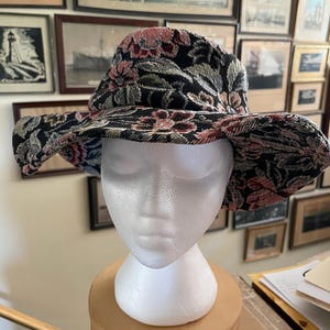 May include: A bucket hat featuring a black base with a woven floral pattern in shades of pink, green, and cream. The hat has a wide, flexible brim and a rounded crown, constructed from a textured fabric that displays a detailed botanical design.