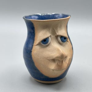 May include: A handmade ceramic vase with a face design. The vase has a blue rim and exterior, with a tan face sculpted onto the front. The face has blue eyes and a carved mouth, giving it a unique, expressive appearance.