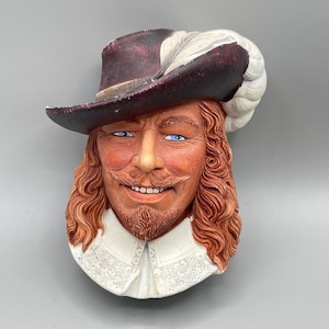 May include: A wall-mounted decorative plaque featuring a sculpted bust of a person with long wavy reddish-brown hair, a mustache, and a goatee. The figure wears a dark maroon wide-brimmed hat with a large white feather and a white lace collar.