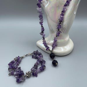 May include: A necklace and bracelet set featuring amethyst chip beads. The necklace has a pendant with two dark purple beads. The bracelet has a silver-tone flower clasp. The jewelry is displayed on a white, pearlescent decorative object.