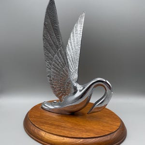 Può includere: Una scultura a forma di cigno cromato con ali spiegate, montata su una base ovale in legno. Le ali del cigno sono decorate con motivi di piume. La base è di colore marrone caldo.