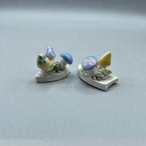 May include: Two miniature ceramic boats, each featuring a cluster of colourful mushrooms. The mushrooms are painted in shades of blue, yellow, and purple. The boats are white and have a rough, textured surface.