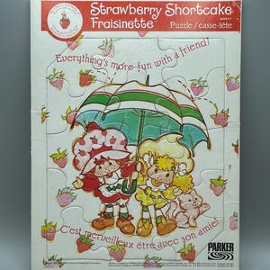 May include: A vintage Strawberry Shortcake puzzle box. The box features the text "Strawberry Shortcake Fraisinette" and "Everything's more fun with a friend!" The puzzle shows two cartoon characters under an umbrella, surrounded by strawberries.
