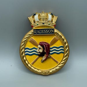 May include: A gold-colored Radisson hotel emblem with a yellow center featuring a man in a red hat, crossed oars, and blue waves. The emblem has a crown at the top and the word "RADISSON" in a blue rectangle.