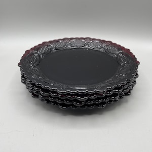 May include: A stack of four deep red glass plates with a smooth, flat centre and a decorative scalloped rim featuring a raised, textured pattern of repeating oval shapes and small dots around the entire circumference of each plate.