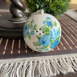 Puede incluir: Un adorno de cerámica blanca con un diseño floral azul y verde. El adorno tiene un agujero en la parte superior para colgar.