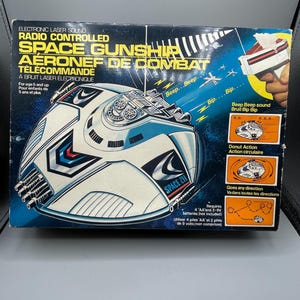 Op de afbeelding: Vintage Space Gunship speelgoeddoos. De doos heeft een futuristisch ruimteschipontwerp in wit, blauw en rood, met de tekst "SPACE GUNSHIP" en "AERONEF DE COMBAT". Het bevat details over elektronisch lasergeluid en radiografische besturing.