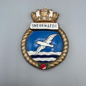 May include: An emblem with a white seagull flying over blue water, framed by a gold-coloured rope border. The emblem includes the word "SHEARWATER" and a crown.
