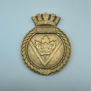 May include: A gold-coloured metal badge featuring the word "VENTURE" above a V-shaped design and a maple leaf. The badge has a rope-like border and is topped with a crown.