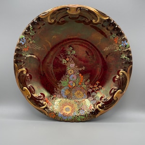 May include: An antique decorative plate with a deep red center and a gold-coloured, ornate rim. The plate features a floral design in the center and along the rim, with accents of blue, orange, and green. The plate is approximately 25 cm in diameter.