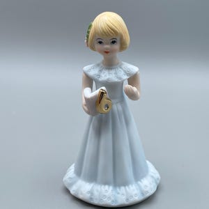 Pode incluir: Estatueta de porcelana de uma menina com um vestido azul claro, segurando o número 6 dourado. A estatueta tem cabelo loiro e um acessório floral para o cabelo. O vestido tem gola e bainha com babados.