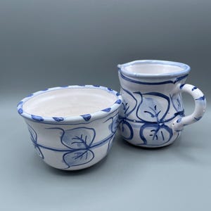 Puede incluir: Un juego de cuenco y jarrita de cerámica blanca. Ambas piezas están decoradas con motivos y ribetes de hojas azules. El cuenco tiene un borde festoneado. La jarrita tiene un asa y un pico. El juego es probablemente para servir o exhibir.