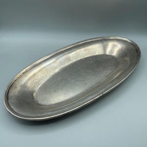 May include: An antique, oval-shaped silver-coloured metal serving tray. The tray has a slightly textured surface and a raised rim. A small, embossed design is visible near one end. The tray is empty and appears to be in good condition.