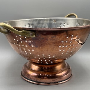 May include: A round copper colander featuring a polished metallic interior, a wide pedestal base, and two curved brass handles attached to the sides. The exterior surface displays a warm, weathered copper finish with numerous drainage holes.