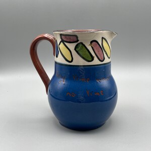 Op de afbeelding: Een blauwe keramische kan met een bruine handgreep. De kan heeft een witte band met kleurrijke abstracte vormen en de tekst "Only time means no time" in bruin.