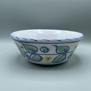Puede incluir: Cuenco de cerámica blanca con borde azul y diseño floral pintado a mano. El cuenco presenta hojas verdes y acentos de flores amarillas, con detalles azules en forma de vid. El cuenco es de tamaño mediano, adecuado para servir o exhibir.