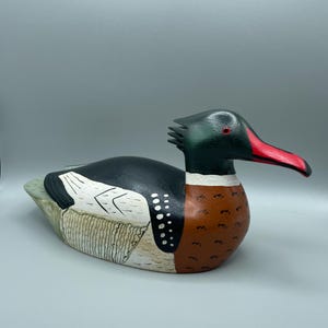 Puede incluir: Escultura de pato de madera tallada a mano. El pato presenta una cabeza y espalda negras, un pecho marrón y un pico rojo. Marcas blancas y negras en el lateral. La escultura está pintada con un acabado mate y tiene un aspecto texturizado detallado.