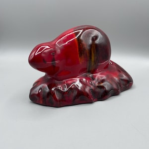 Puede incluir: Una figurita de cerámica roja y marrón de un ratón sentado sobre una base. El ratón está enroscado y tiene un acabado brillante.