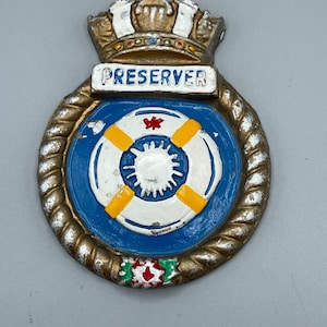 Może przedstawiać: Metalowa odznaka w stylu vintage z koroną i napisem "PRESERVER". Odznaka przedstawia niebiesko-biały wzór koła ratunkowego z żółtymi akcentami, otoczony złotą obwódką z liny. Czerwony kwiatowy wzór znajduje się na dole.