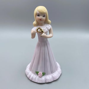 May include: A porcelain figurine of a young girl with blonde hair, wearing a long, light pink dress. She holds a gold number 9. A pink rose and green leaves adorn the base. A collectible birthday cake topper.