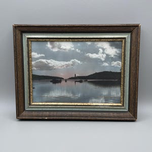May include: Framed landscape art depicting a tranquil coastal scene. The artwork shows a body of water with a distant shoreline, under a cloudy sky. The frame is dark brown with a light blue and gold inner border.