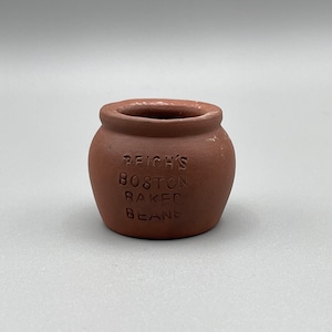 May include: A small, round, terracotta-colored pot with a wide opening. The words "REICH'S BOSTON BAKED BEANS" are embossed on the front. The pot has a slightly rough texture and a simple, rustic design.