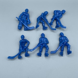 Op de afbeelding: Zes kleine, blauwe plastic hockey speler beeldjes. Elke figuur staat in een andere pose, met een hockeystick. De beeldjes zijn gedetailleerd met uitrusting en kleding, tegen een lichtgrijze achtergrond.