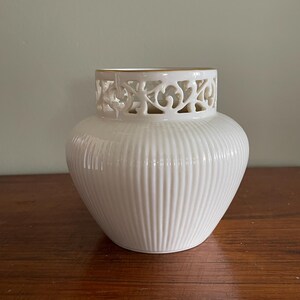 May include: A cream-colored ceramic vase with a rounded, ribbed body and a decorative openwork collar featuring intricate cut-out scroll patterns. The top rim of the vase is finished with a thin, metallic gold band, providing a subtle contrast.