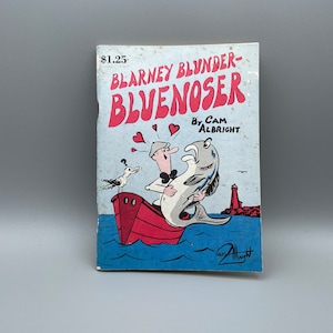 Op de afbeelding: Een vintage stripboek getiteld "Blarney Blunder-Bluenoser" door Cam Albright. De cover toont een cartoonillustratie van een man in een boot met een vis, een zeemeeuw en een vuurtoren. De titel is rood en de achtergrond is blauw.