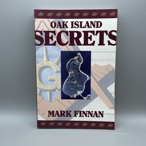 May include: A hardcover book titled "Oak Island Secrets" by Mark Finnan. The cover features a map of an island, a Masonic symbol, and a decorative border with leaf patterns. The title and author's name are in maroon.
