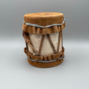 May include: A small, brown leather-covered drum with a birch bark body. The drum has a decorative pattern of brown leather strips and a blue leather trim.