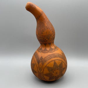Può includere: Una zucca artigianale con un collo lungo e curvo e una base arrotondata. La zucca è di un caldo colore arancione ed è decorata con intricati motivi incisi marrone scuro, tra cui forme geometriche e motivi floreali. La zucca è esposta su uno sfondo grigio.