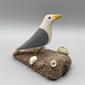 May include: A handcrafted seagull figurine with a white head, grey wings, and a yellow beak with a red eye detail. The bird stands on a textured, sand-covered rectangular base decorated with a small white seashell and a green sea urchin shell.