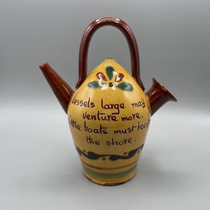 May include: A yellow ceramic vessel with a brown handle, spout, and rim. The body features a hand-painted floral motif and the text: Vessels large may venture more. Little boats must keep the shore. It has green and brown horizontal bands.