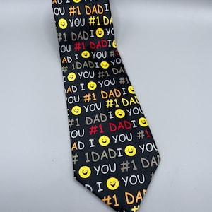 May include: Black necktie with a repeating pattern of the text "#1 DAD" in white, red, and brown, along with yellow smiley faces. The words "I YOU" are also included in the design.