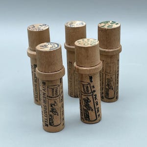 Könnte beinhalten: Eine Sammlung von Vintage-Holzspulen mit aufgedrucktem Text und Grafiken. Die Spulen sind zylindrisch mit breiterem Boden und schmalerem Oberteil. Der Text enthält den Markennamen "Crafts Brand" und den Satz "Buy New Shuttle".