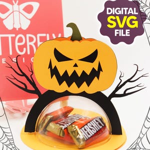 Puede incluir: Un portador de caramelos con temática de Halloween con una calabaza de papel con una cara tallada. La calabaza se asienta sobre una silueta negra con ramas, y una cúpula transparente contiene barras de chocolate Hershey's. Las palabras "DIGITAL SVG FILE" están en una burbuja morada.
