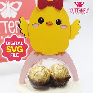 Puede incluir: Una caja de regalo amarilla con forma de pollito con un lazo rojo, que contiene dos bombones dorados. El diseño incluye el texto "DIGITAL SVG FILE" y el logotipo "CUTTERFLY DESIGN". La caja está sobre una base rosa y blanca.