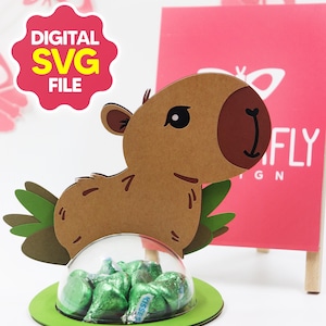Op de afbeelding: Een bruin, capibara-vormig papieren ambacht met een doorzichtige koepel met groen verpakte snoepjes. Het ambacht heeft een groene basis en staat voor een roze bord met het woord "FLY" en de tekst "DIGITAL SVG FILE".