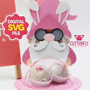 Puede incluir: Un gnomo de papel rosa con orejas de conejo, gafas de sol y barba blanca, sosteniendo dos golosinas envueltas. La imagen incluye el texto "DIGITAL SVG FILE" y "CUTTERFLY DESIGN". El gnomo está sobre una base circular rosa.