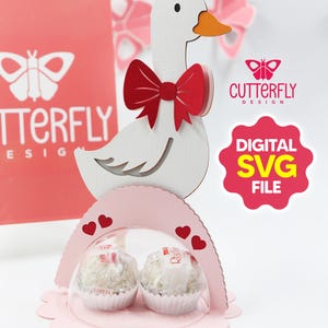 Puede incluir: Una manualidad de papel con forma de ganso blanco con pajarita roja, que contiene dos caramelos envueltos en una cúpula transparente. El diseño incluye una base rosa festoneada y detalles de corazón. También aparece el texto "DIGITAL SVG FILE".