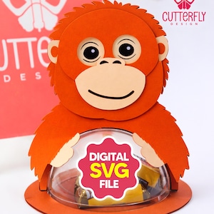 Puede incluir: Orangután de papel naranja con una cúpula transparente que contiene golosinas. El orangután tiene una cara sonriente, ojos negros y un hocico de color beige claro. La base tiene el texto "DIGITAL SVG FILE". El fondo tiene el texto "CUTTERFLY DESIGN".
