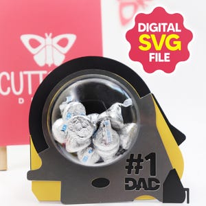 Puede incluir: Un portador de caramelos en forma de cinta métrica negra, amarilla y gris, lleno de caramelos de chocolate envueltos en plata. El portador tiene el texto "#1 DAD" recortado en la parte delantera. Una tarjeta rosa con un diseño de mariposa está en el fondo. La imagen también incluye una etiqueta roja que dice "DIGITAL SVG FILE".