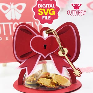 Puede incluir: Una caja de regalo roja en forma de lazo, con un diseño de cerradura y llave en forma de corazón. Contiene una cúpula de cristal transparente llena de monedas de chocolate doradas. Las palabras "DIGITAL SVG FILE" están en una burbuja rosa.