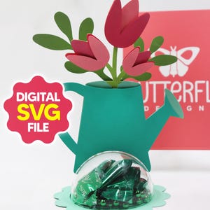 Puede incluir: Una regadera de papel turquesa con tulipanes de papel rosa y hojas verdes. Una cúpula de plástico transparente llena de caramelos verdes envueltos se asienta sobre una base verde festoneada. Las palabras "DIGITAL SVG FILE" están en una estrella roja.