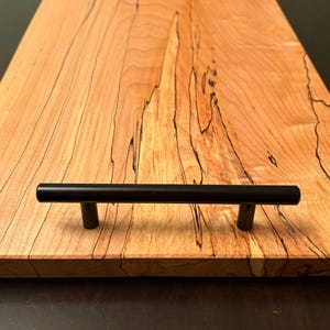 May include: A rectangular wooden serving tray with a light brown finish and dark grain patterns. The tray has two black metal handles. The tray is on a dark surface.