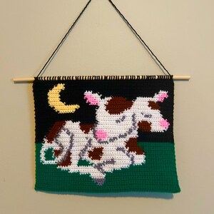 Pode incluir: Uma tapeçaria de parede de crochê que representa uma vaca branca com manchas marrons dormindo sob uma lua crescente amarela em um fundo preto. A vaca está deitada na grama verde.